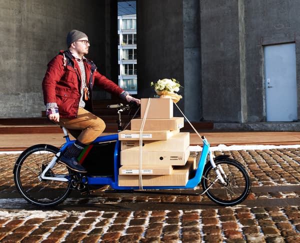 An customer transporting flatpacks by a long bicycle across a cobblestone sidewalk.