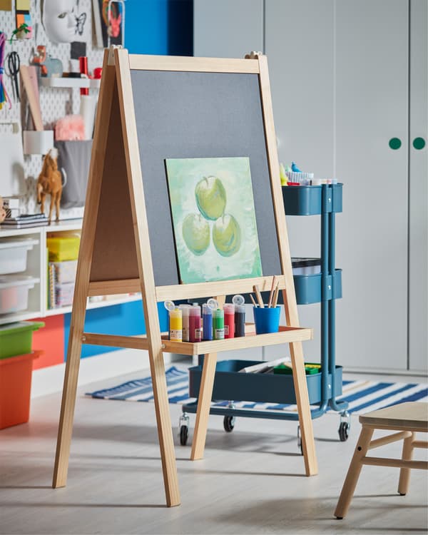 An easel in softwood that has an included tray where paint in different colors stands.
