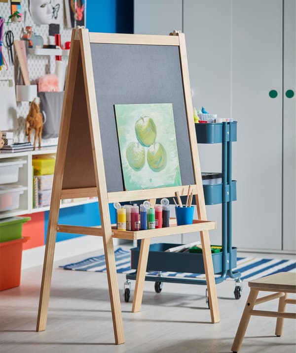 An easel in softwood that has an included tray where paint in different colours stands.