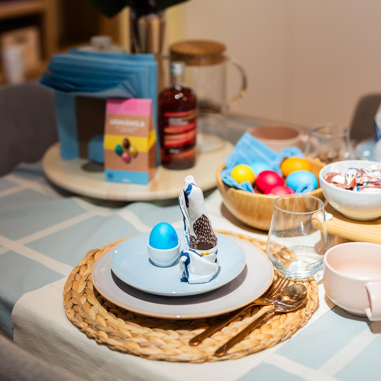 An Easter table setting dominated by light blue and soft pink hues. A woven place mat, plates, and a bright blue egg in a cup. In the background a wooden bowl with brightly colored eggs, a package of chocolate treats, and a bottle of syrup on a rotating wooden tray.