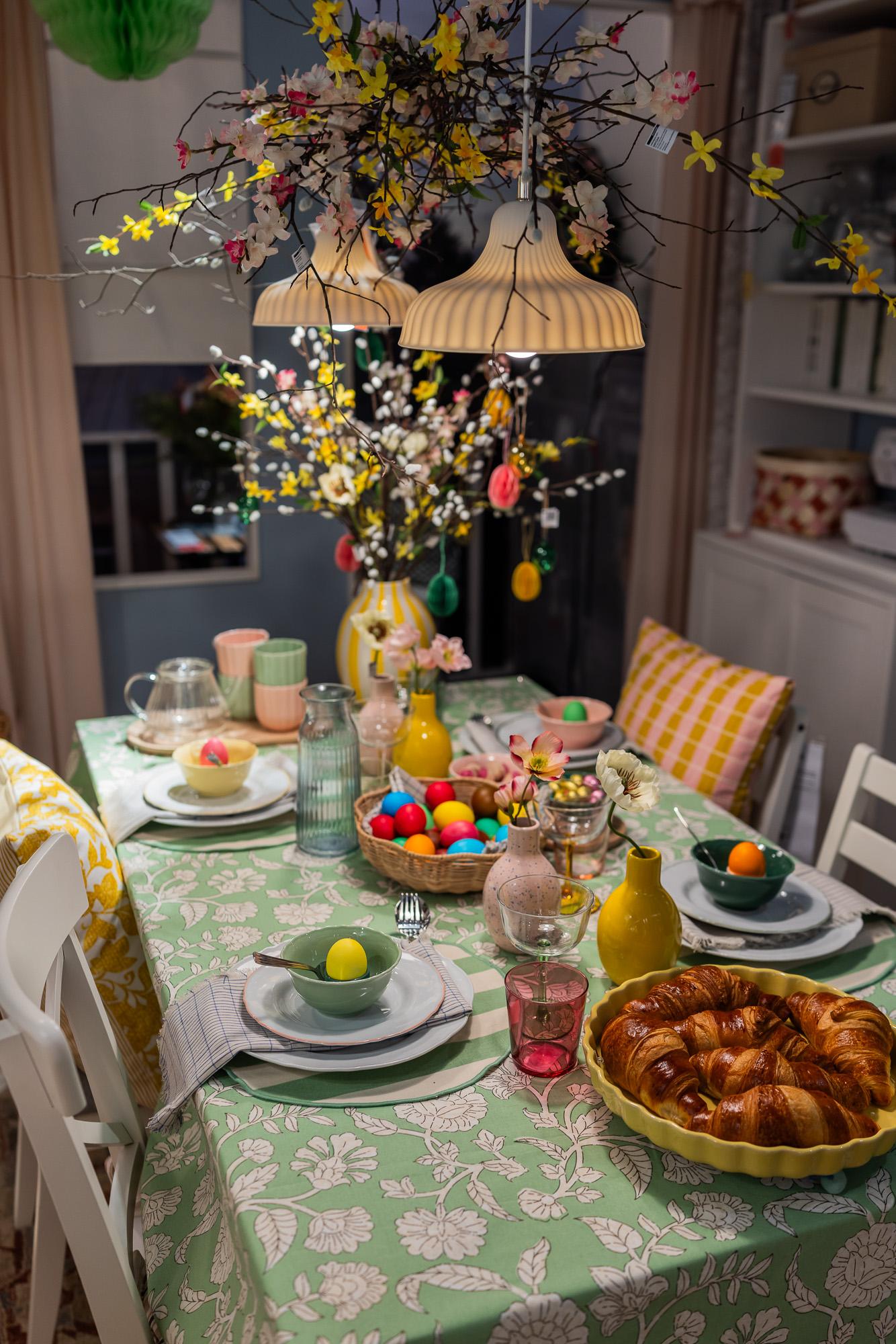 An Easter table setting featuring a green and white tablecloth with a floral pattern, colored eggs in the center of the table, and a bouquet of branches in a vase at the corner of the table. Two pendant lamps with a decorative arrangement of branches between them hanging above the table.