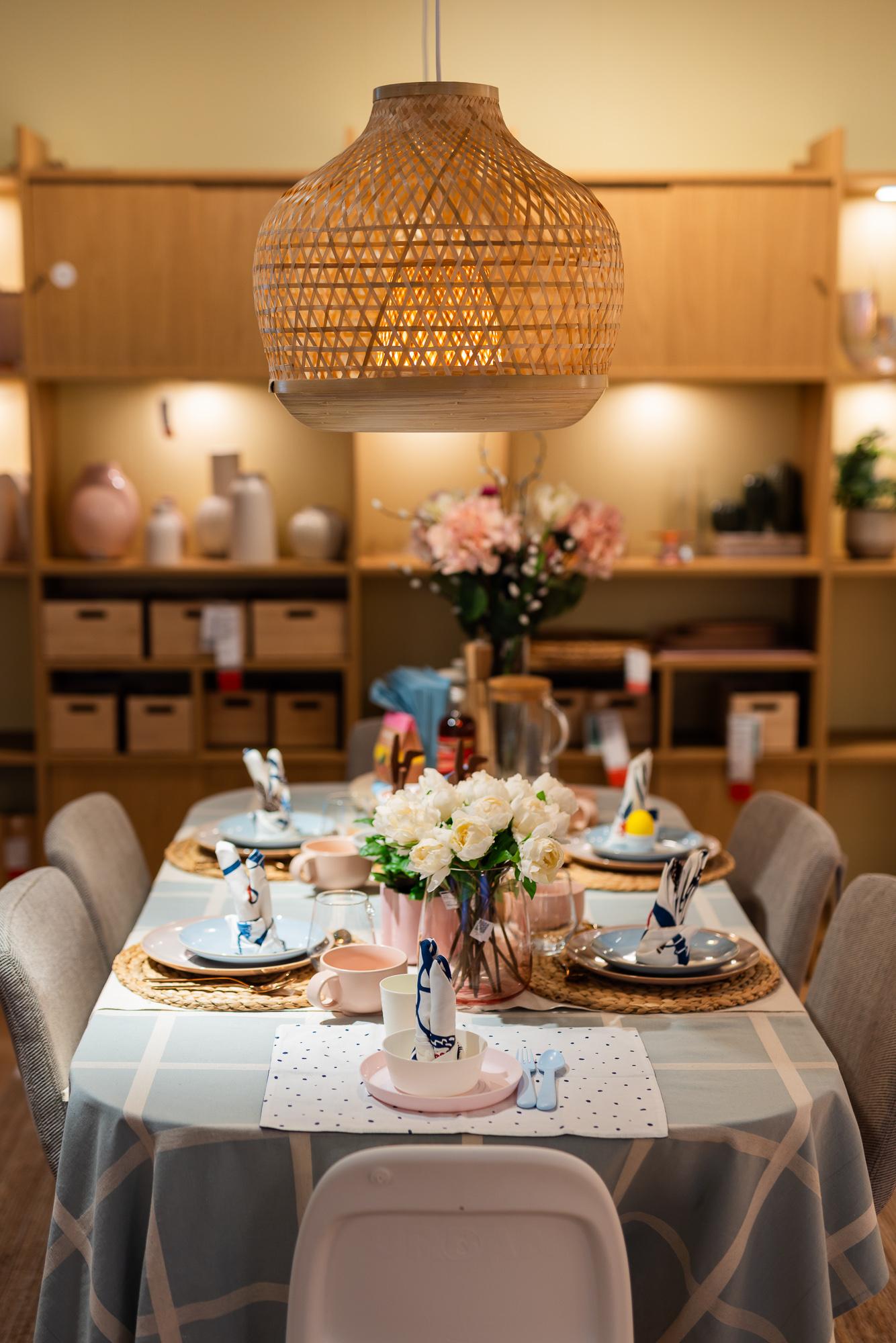 An Easter table setting in a cozy living room with a large, woven lampshade hanging above the table. On the table: plates in soft shades of pink and blue, decoratively folded napkins, and flowers in vases.