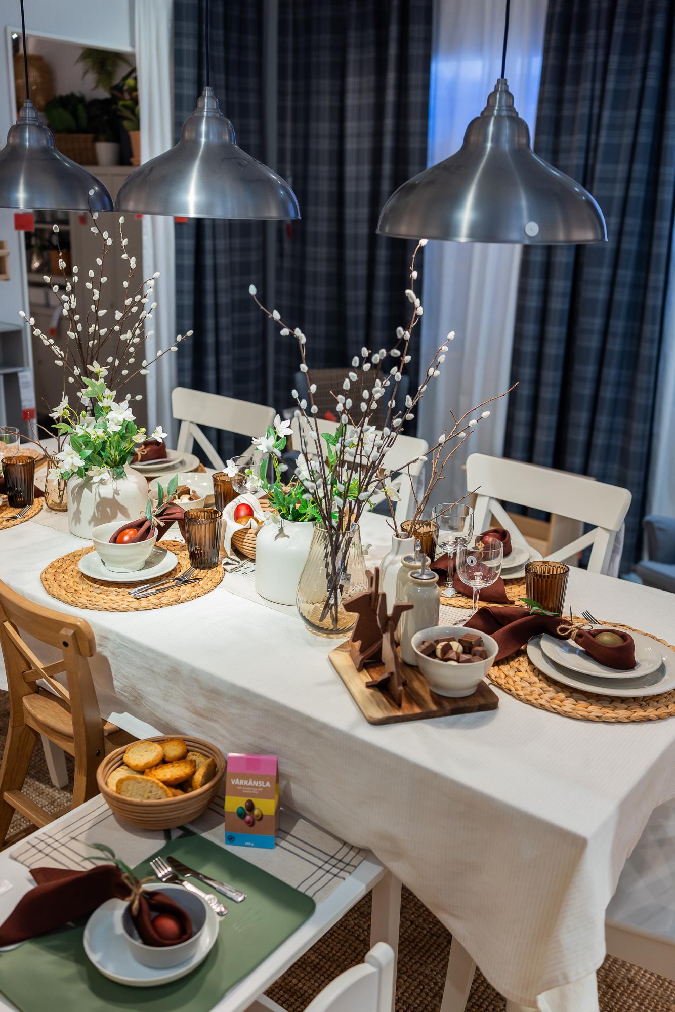 An Easter table setting in the living room featuring a white tablecloth, woven place mats, light-coloured plates, and willow arrangements in vases; a smaller beautifully set table nearby for a child.
