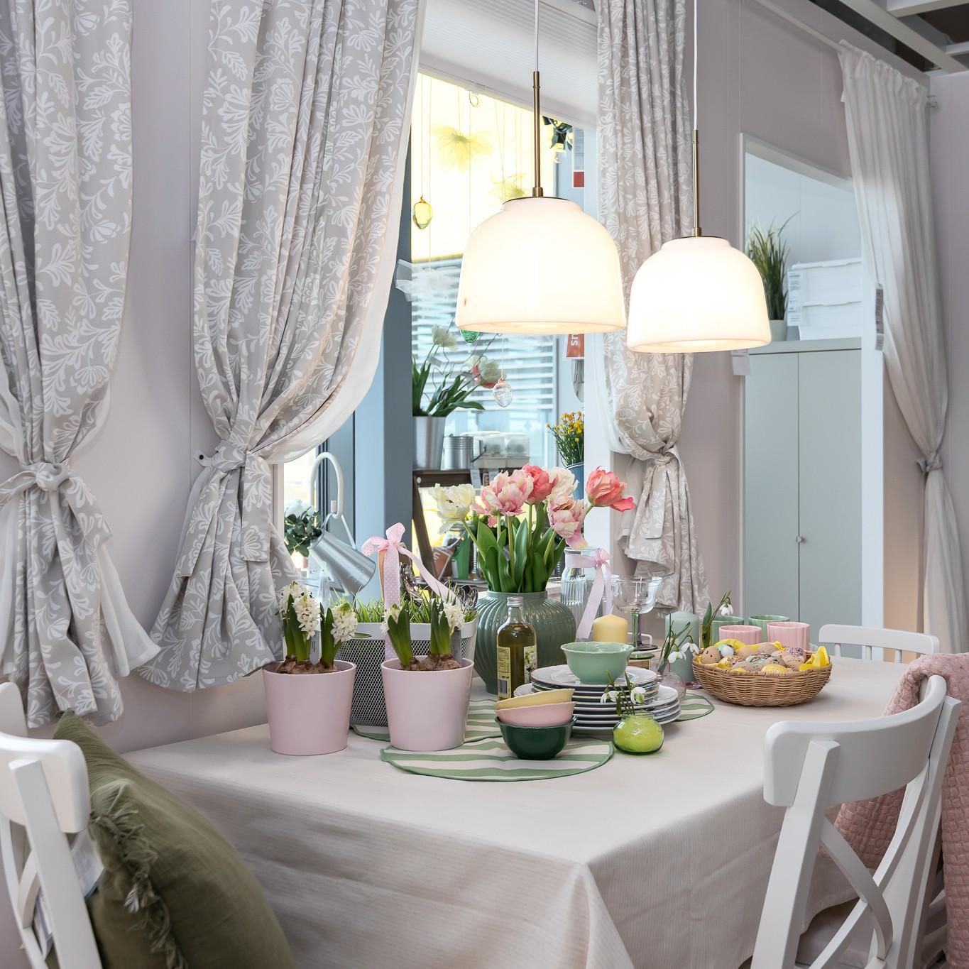 An Easter‑themed kitchen table decorated with colourful tulips, potted plants, pastel candles and tableware, set near large windows with light curtains.