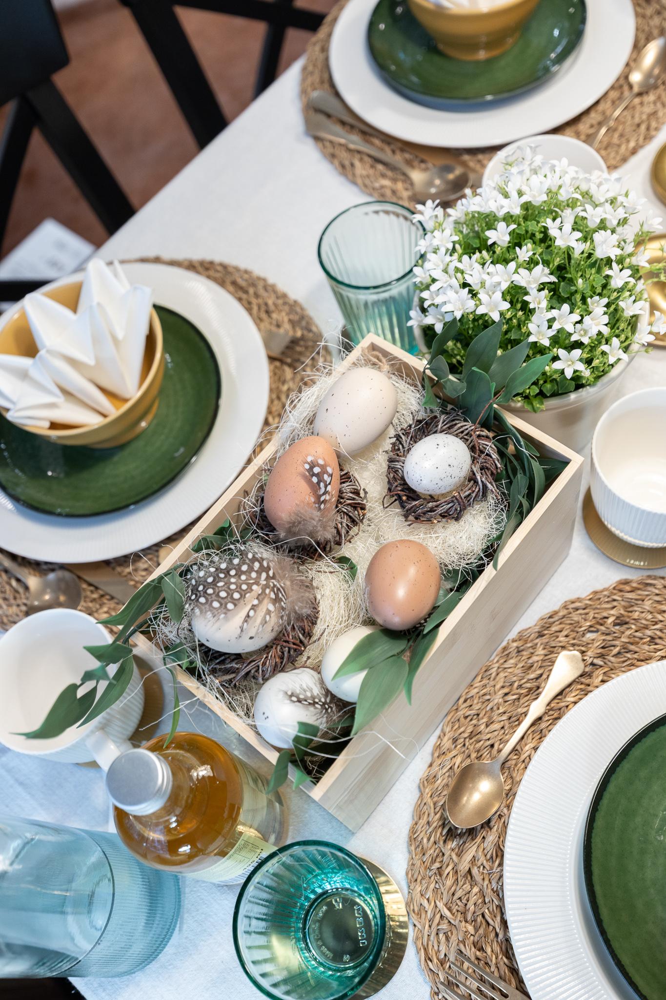 An Easter-themed table set with layered green and white plates, yellow bowls, folded napkins, tall candles, glassware, and small white floral arrangements on a white tablecloth.