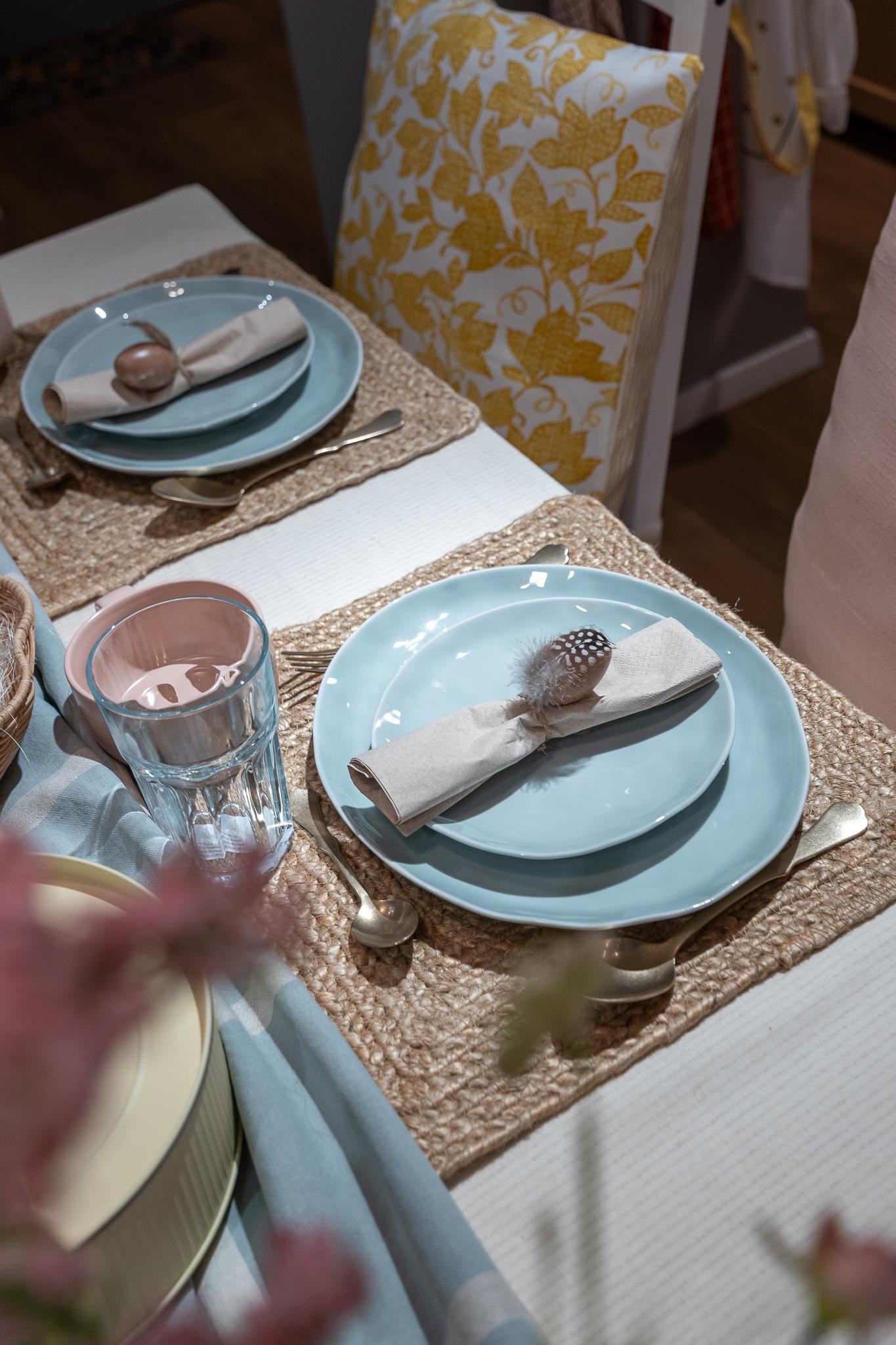 An Easter-themed table setting featuring pastel blue plates, woven placemats, a bowl of decorative Easter eggs, candles, and colourful tableware.