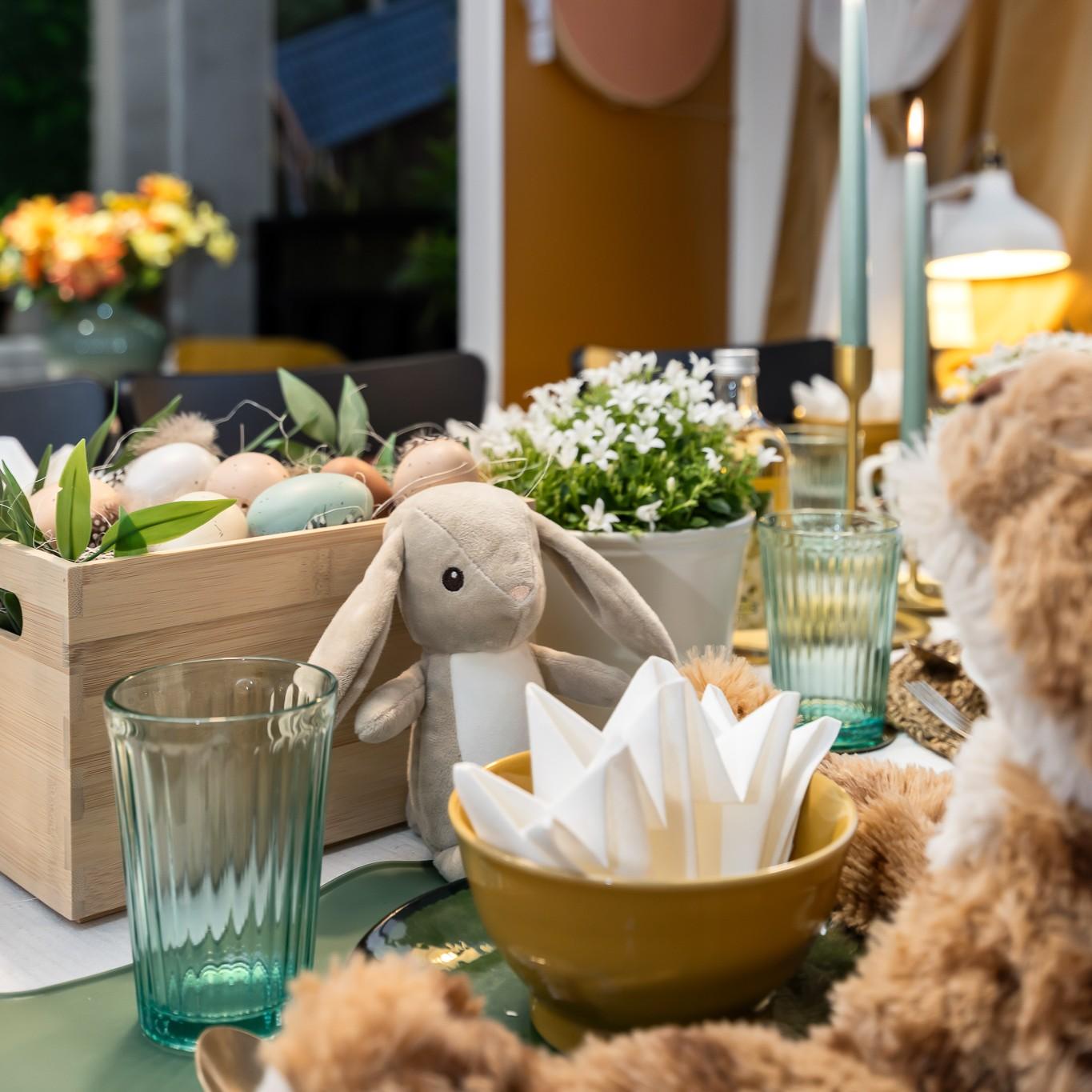 An Easter-themed table with green-tinted glassware, a wooden box filled with decorative eggs, small white flowers and a bowl with a folded napkin, with soft toys placed around the setting.