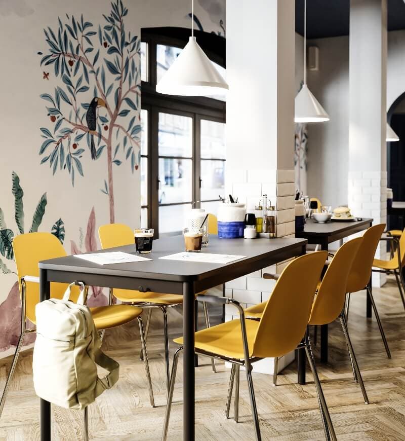 An eatery showcasing white walls with painted trees featuring two black long tables and yellow chairs on a light birch floor.