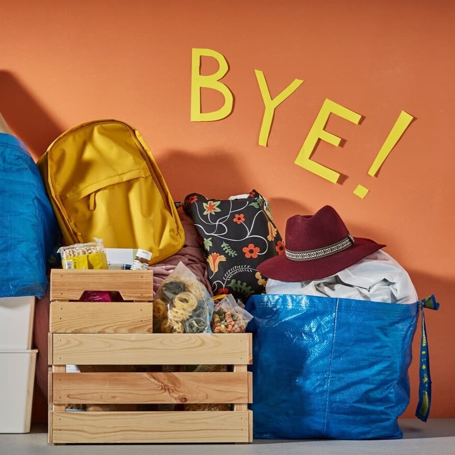 An einer orangenen Wand steht eine Holzkiste zwischen 2 blauen IKEA Taschen.