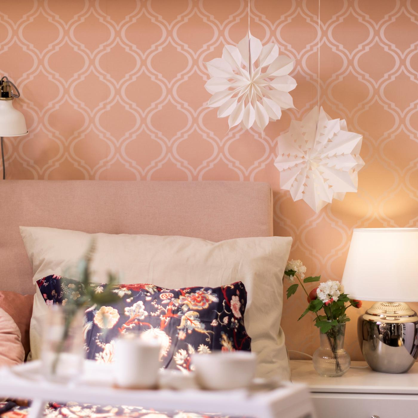 An einer rosanen Wand mit einem weißen Muster steht ein Bett mit einem hellen Kissen und einem nunten mit einem Blumen Muster. Auf der Seite steht ein Nachttisch mit einer Tischlampe darauf.