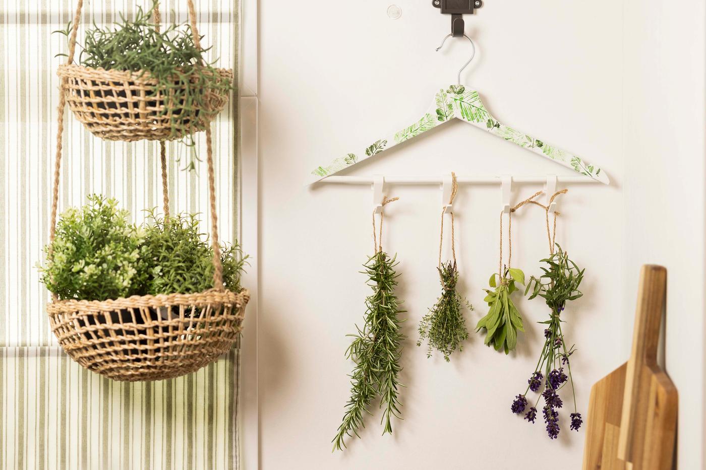 An einer weißen Wand hängt ein weißer Kleiderbügel mit 4 grünen Zweigen. Auf der linken Seite kann man 2 Körbe mit grüner Deko sehen.