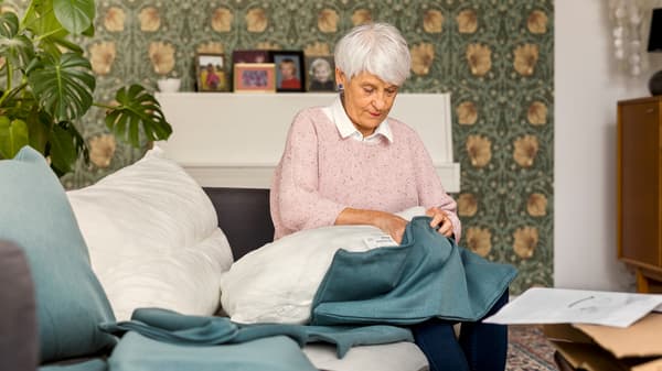 An elderly woman wearing a light pink sweater sits on a sofa while stuffing a sofa cushion in a new blue cover.