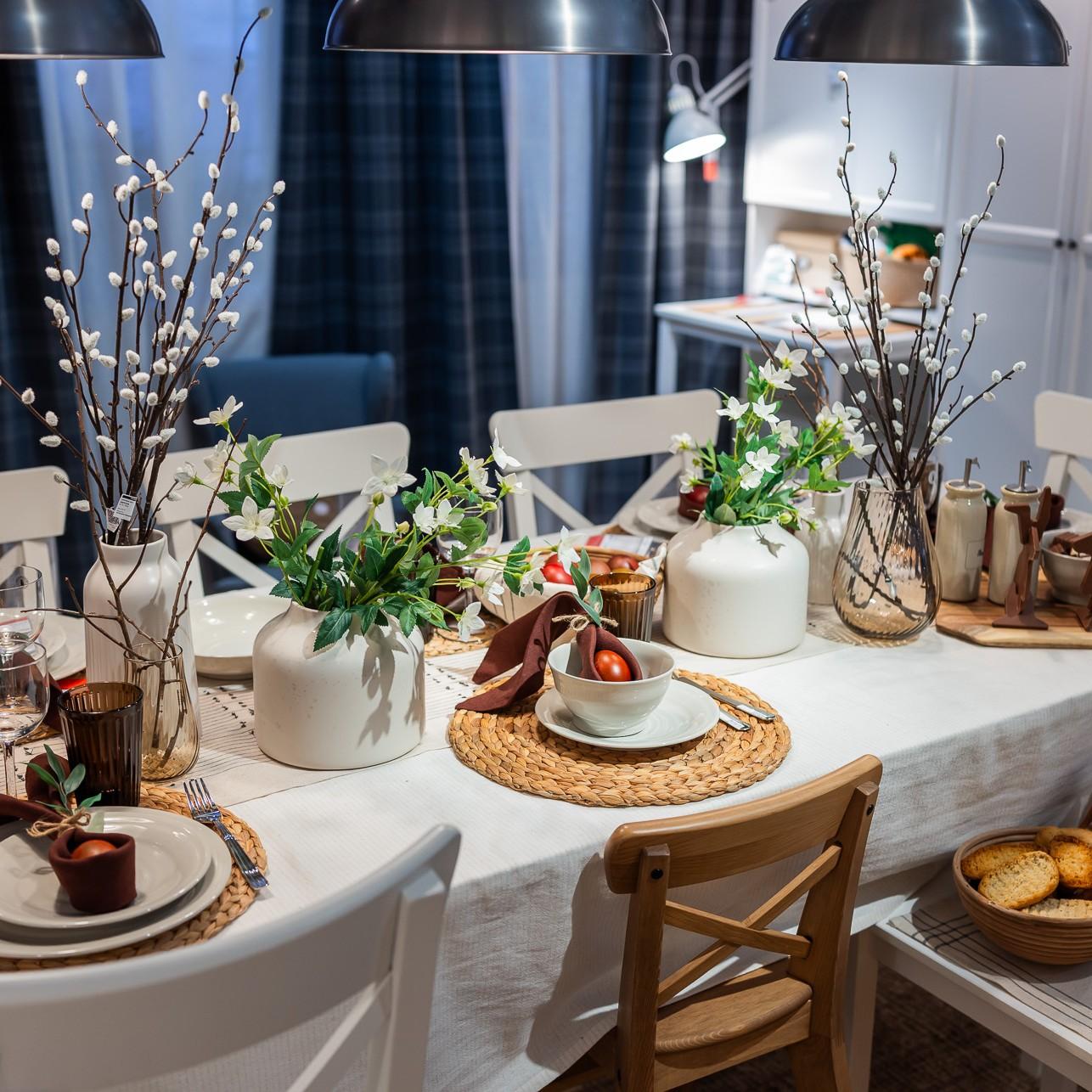 An elegant Easter table setting featuring a white tablecloth, woven place mats under white plates, and white ceramic and tinted glass vases with white flowers and branches in the center of the table.