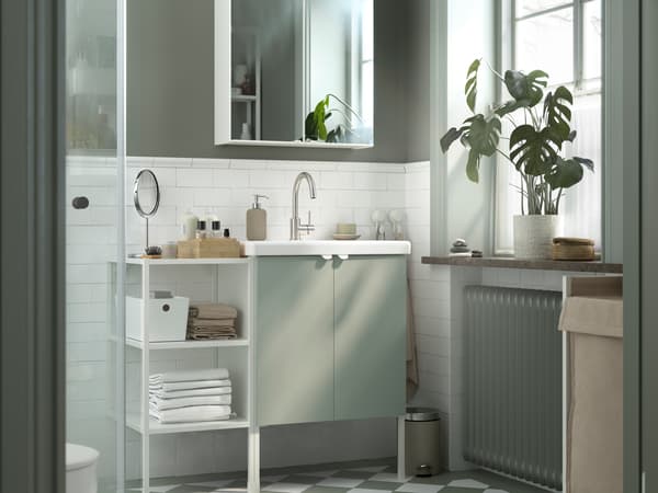 An ENHET cabinet in white with two doors and an open shelf to the left filled with towels, in a grey and white bathroom.