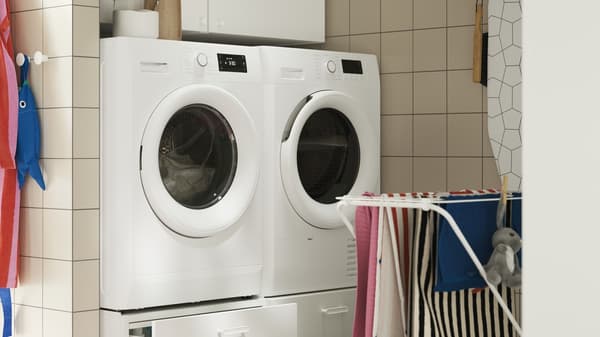 An ENHET pedestal, placed under a washing machine and drier, in a tiled laundry room with a drying rack.
