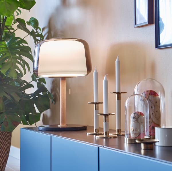 An EVEDAL dome-shaped table lamp next to three GLITTRIG white and gold coloured candle sticks and BEGÅVNING glass domes.