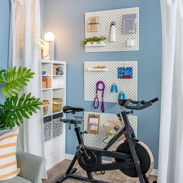 An exercise bike is placed behind an open curtain in a living room. Beside it is a KALLAX shelving unit which is against the wall, and three pegboards holding different items are on the other wall.