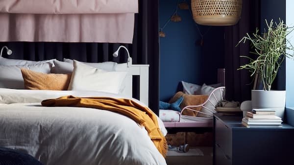 An extendable kids' bed by a white queen bed with beige/white bed linen, a dark beige cushion, pink curtains and a bamboo pendant lamp.