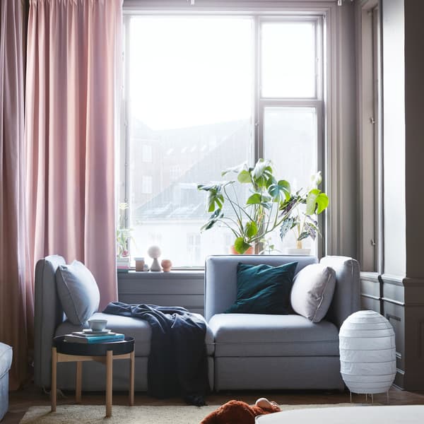An grey IKEA VALLENTUNA 2-seat modular sofa with storage, near a large window with pink curtains.