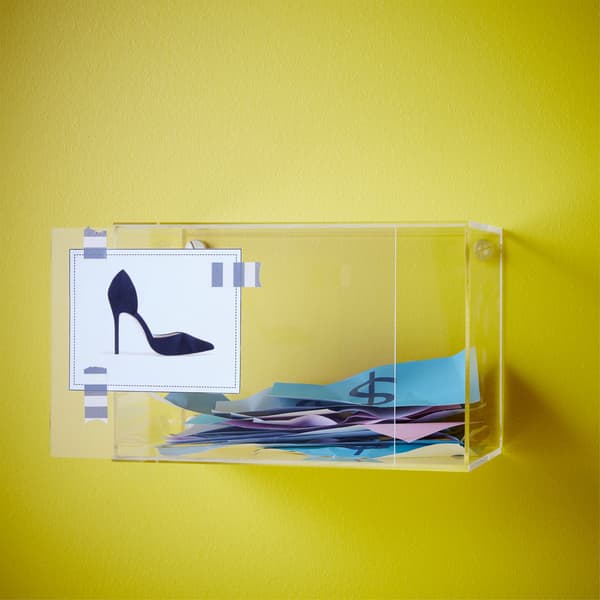 An IKEA BJÖRNARP acrylic display box on the wall with a shoe sketch pinned to the front and pretend money inside.