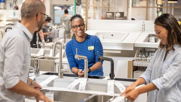 An IKEA co-worker in a blue striped shirt is smiling and talking to two people in-store and showing sinks and faucets