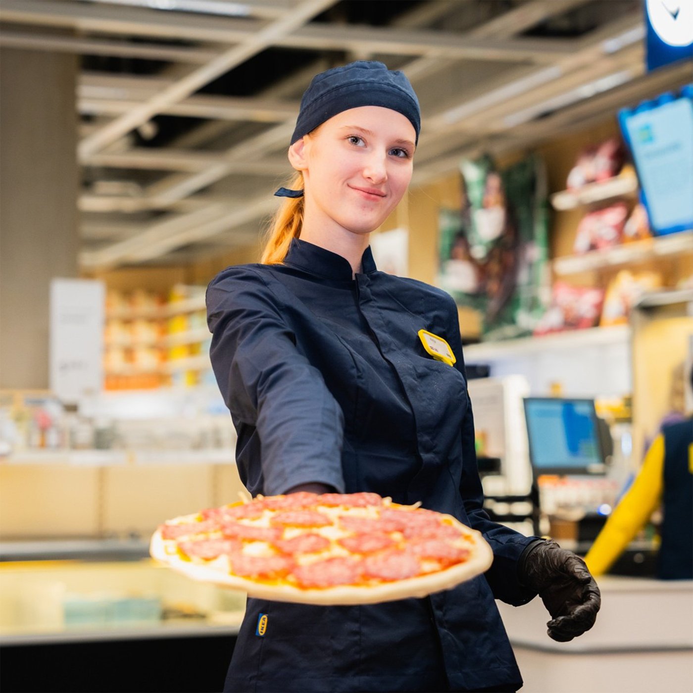 An IKEA co-worker in a dark blue uniform serves a salami pizza on camera, the store layout in the background.