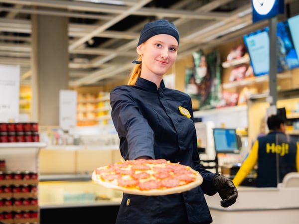 An IKEA co-worker in a dark blue uniform serves a salami pizza on camera, the store layout in the background.