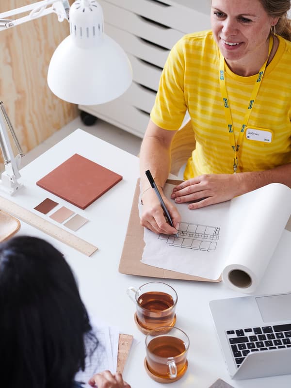 An IKEA co-worker in a yellow top talking to a customer, writing down notes on a plan on a work desk with a white lamp.