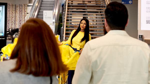 An IKEA co-worker in uniform and carrying a yellow IKEA bag, coming down IKEA store stairs to greet customers.