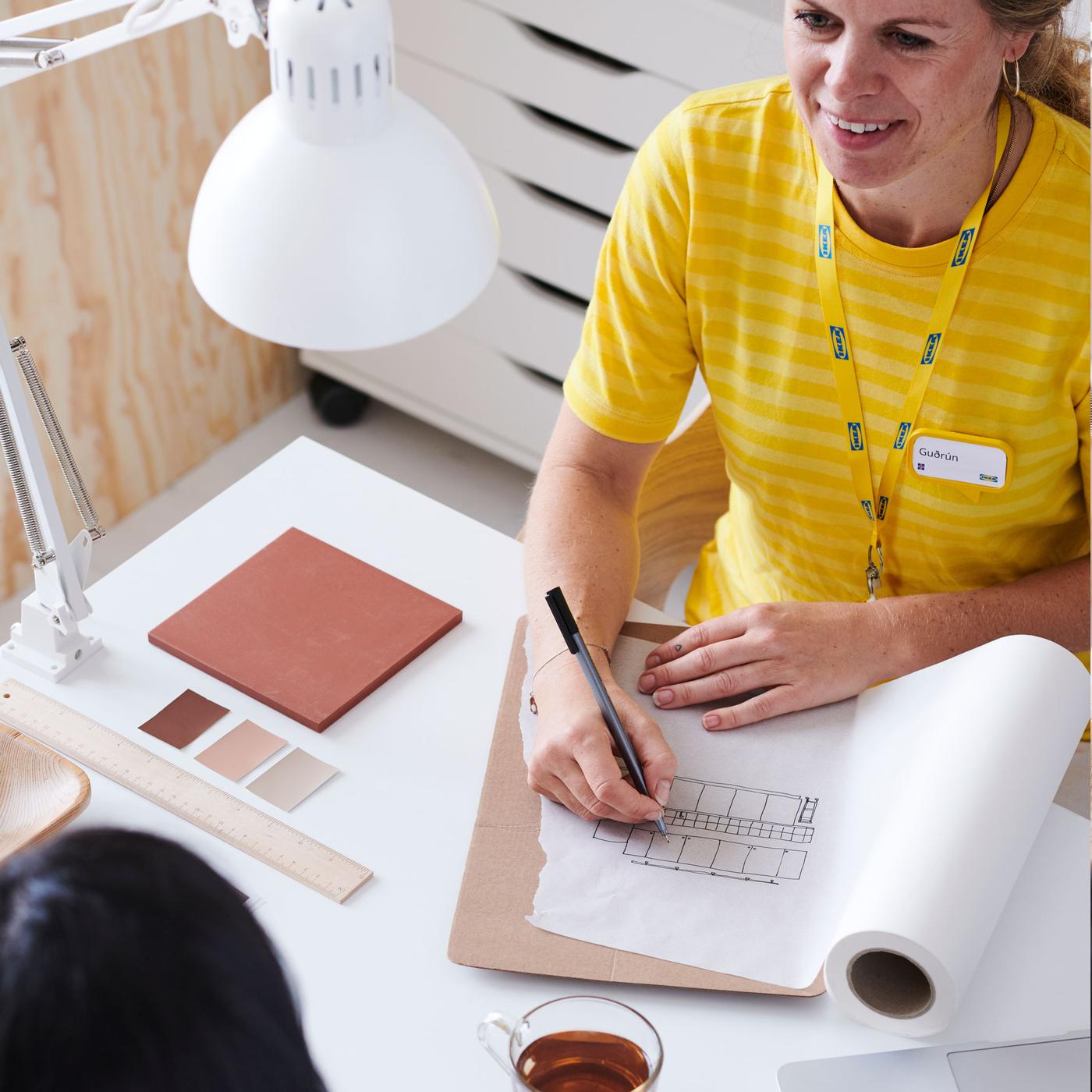 An IKEA co-worker in yellow sitting with a customer drawing plans on a pad of paper.