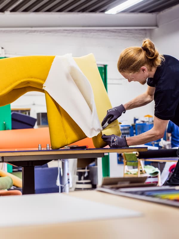 An IKEA co-worker wearing a black t-shirt and work gloves reupholstering an armchair with bright yellow fabric.