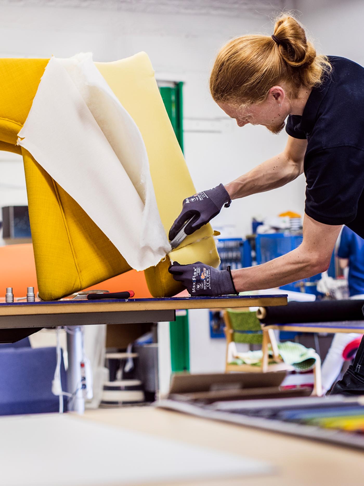 An IKEA co-worker wearing a black t-shirt and work gloves reupholstering an armchair with bright yellow fabric.
