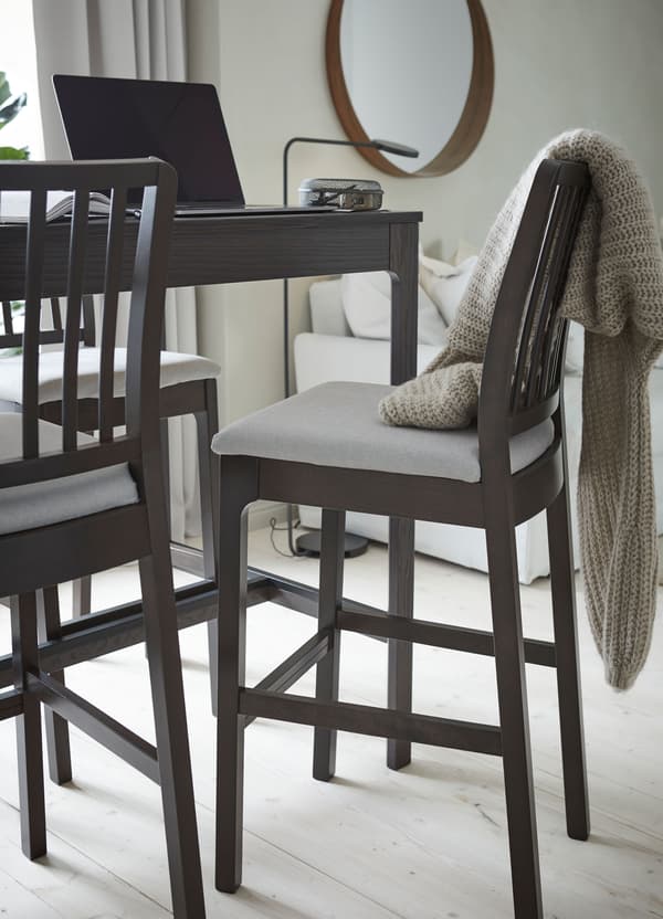 An IKEA EKEDALEN dark brown bar table and bar chairs with light grey cushions, with an open computer on the table.