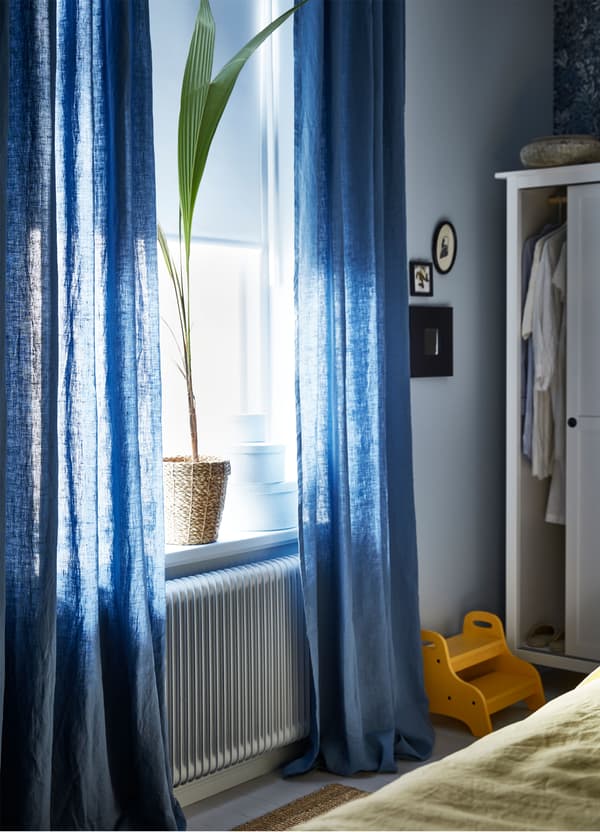 An IKEA FRIDANS white block out roller blind paired with sheer blue AINA curtains, with a large plant on a window sill.