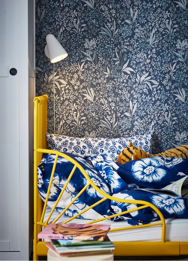 An IKEA FUBBLA white LED wall lamp mounted above a yellow children’s bed, dressed in blue floral SÅNGLÄRKA bedlinen.