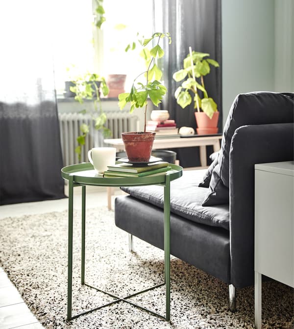 An IKEA GLADOM tray table in dark green with a removable tray top, next to a grey sofa in front of a window.