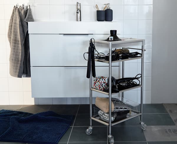 An IKEA GRUNDTAL cart stocked with blowdryers, curling irons, and other accessories stands in front of a bathroom sink.
