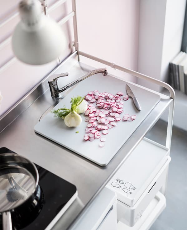 An IKEA GRUNDVATTNET grey plastic cutting board with sliced radish and a knife on top of a sink on a SUNNERSTA kitchenette.