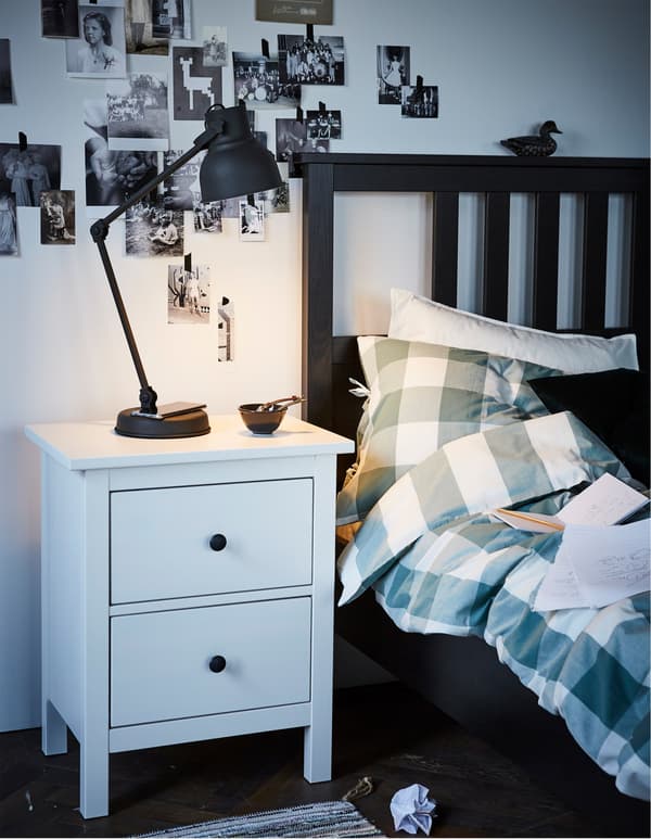 An IKEA HEKTAR work lamp in dark grey with wireless charging on a bedside table next to a bed with checked bed linen.