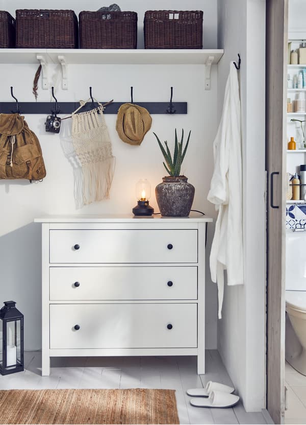 An IKEA HEMNES white chest of 3 drawers in a bedroom, with a rail and shelf above holding baskets.