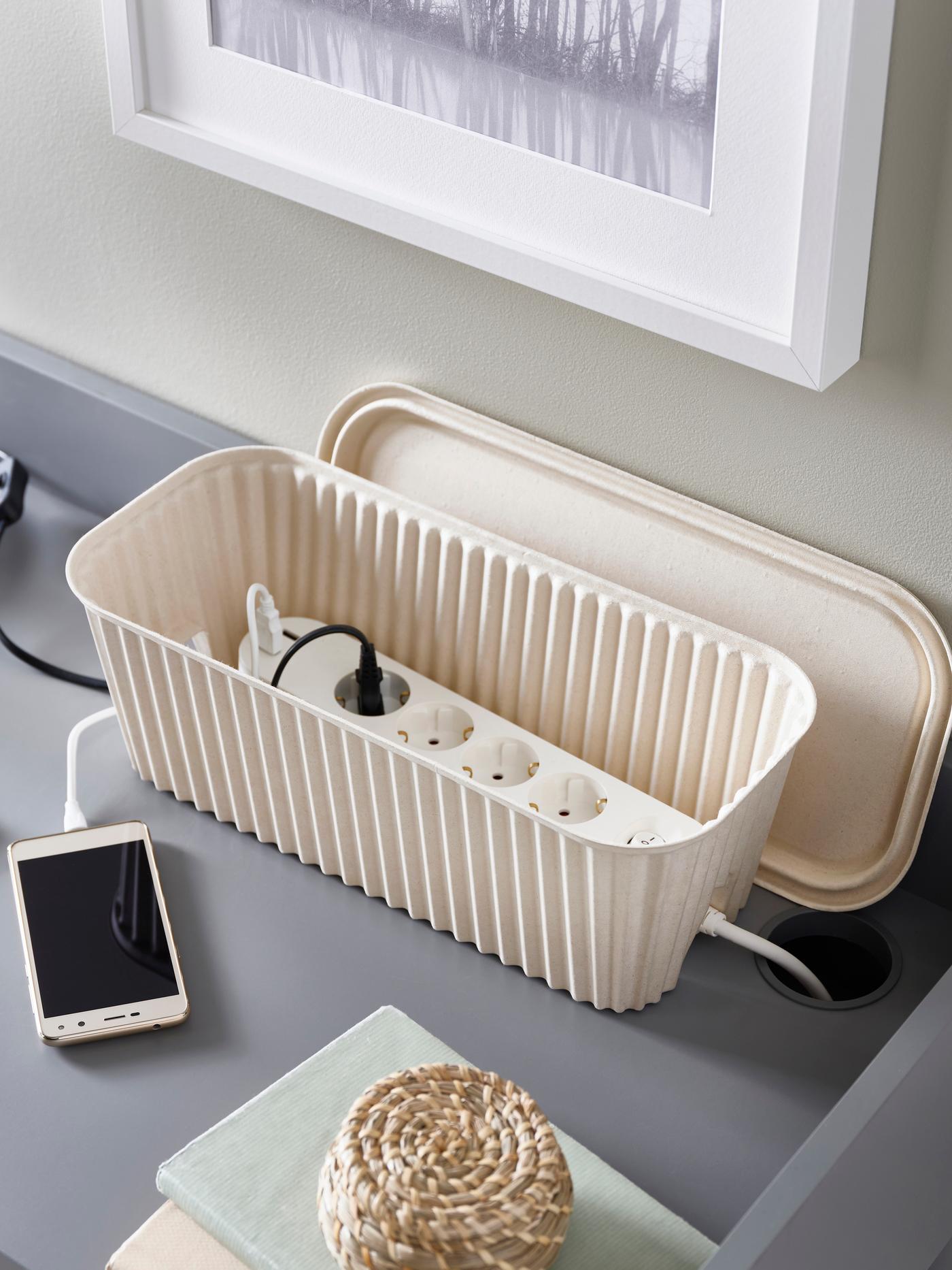 An IKEA HÖNSNÄT cable management box in a natural colour, with a plug extension inside, on a grey desk.