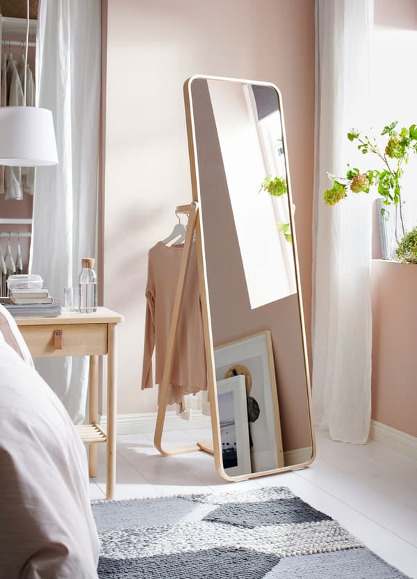 An IKEA IKORNNES long standing dressing mirror positioned near a window, with clothes hanging on the rail behind the mirror.