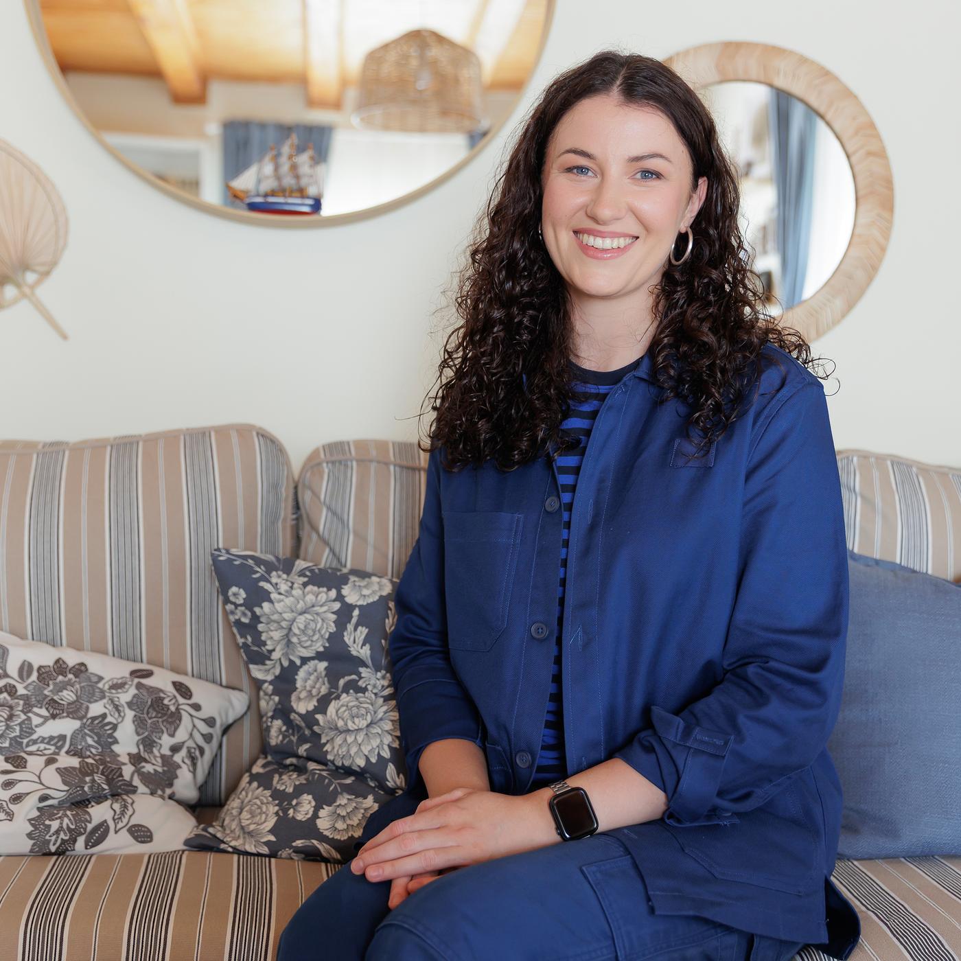 An IKEA interior designer, dressed in a blue uniform, is sitting on a striped sofa-bed, smiling at the camera. They are surrounded by decorative cushions in various shades of blue and floral patterns. Round mirrors and framed artwork are visible in the background.