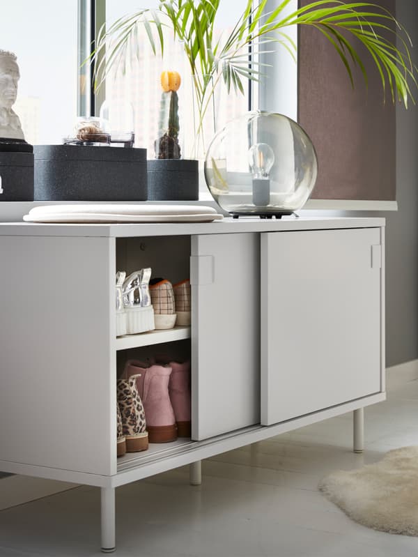 An IKEA MACKAPÄR white shoe bench with storage compartments, sliding doors and a shelf, filled with shoes.
