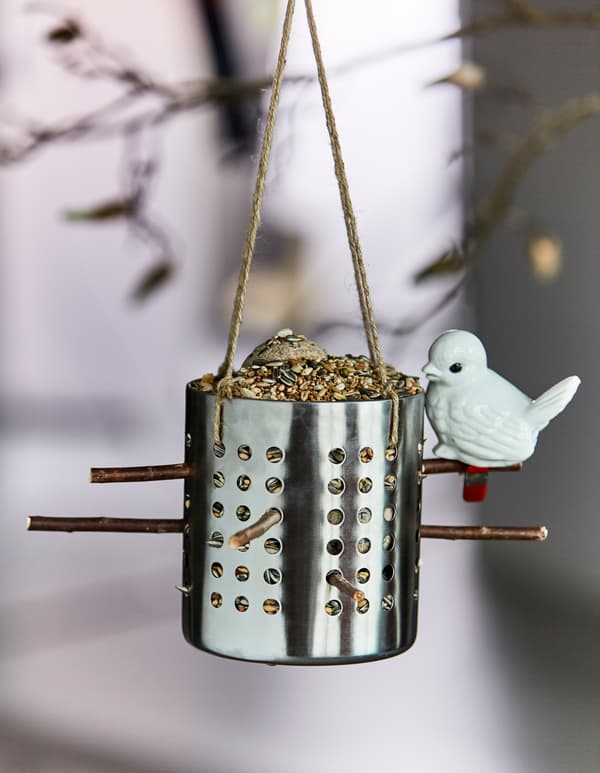 An IKEA ORDNING utensil holder is filled with bird seed and hung to be used as a bird feeder.