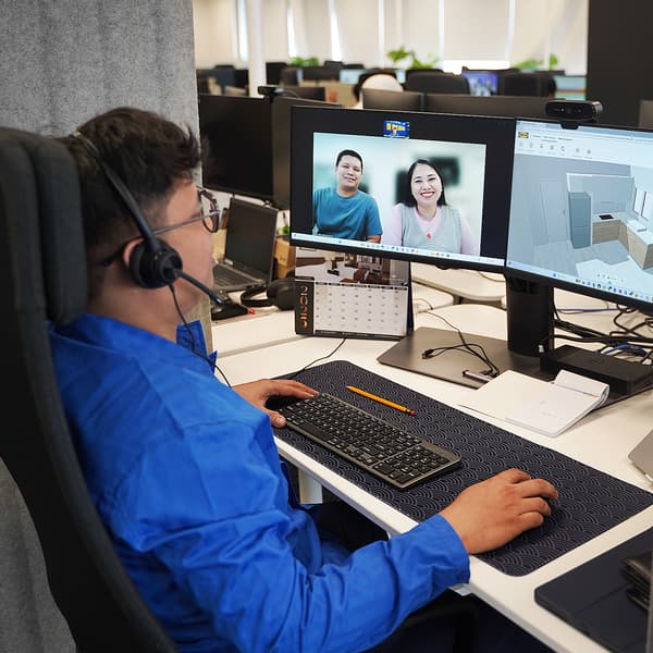 An IKEA Planning Specialist in a headset in a free planning service online session, working at a desk with a computer displaying a video call and a 3D dream home design with an IKEA customer couple. 