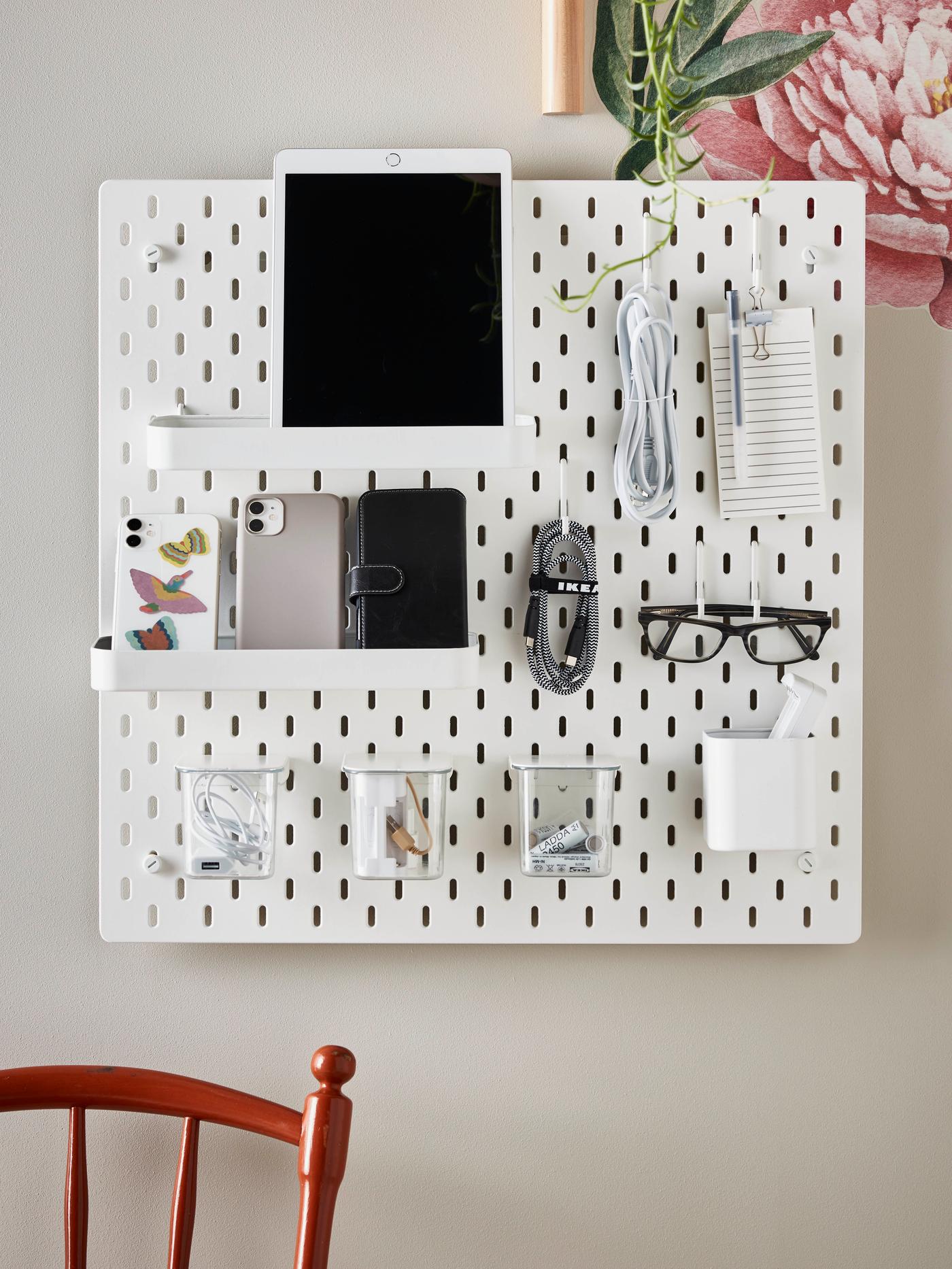 An IKEA SKÅDIS white pegboard on a beige wall with office accessories attached.