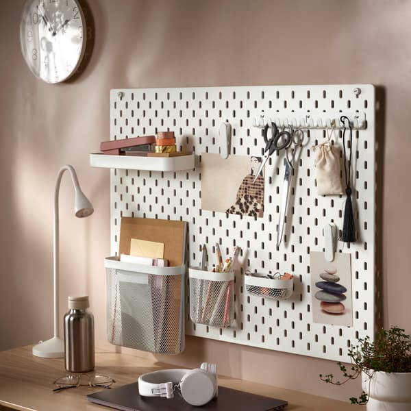 An IKEA SKÅDIS white pegboard with holes holding differently coloured containers storing colourful pens, paper clips and tape.