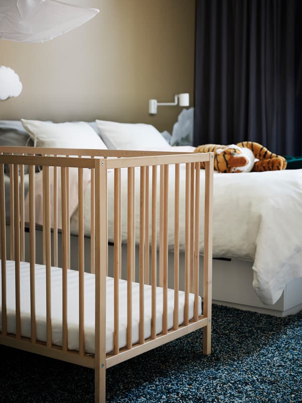 An IKEA SNIGLAR wooden cot in a carpeted bedroom with a double bed and a tiger soft toy and blackout curtains.