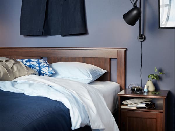 An IKEA SONGESAND brown and sturdy bed frame, beside a bedside table with a SKURUP black work/wall lamp for reading.
